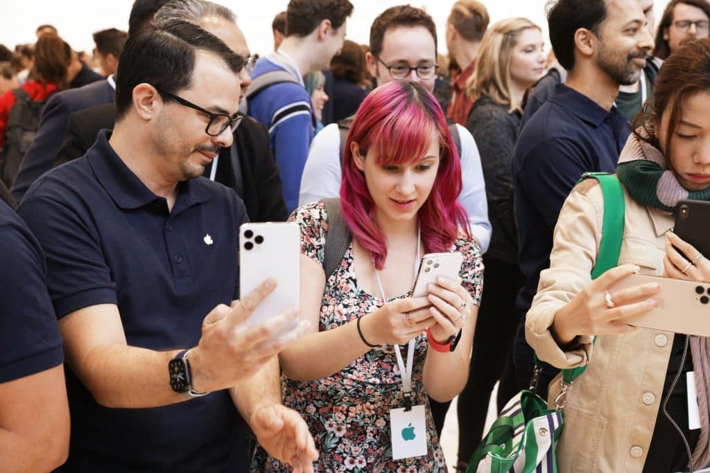 A crowd of people, some holding or using smartphones, gathers at an event. A woman with pink hair and a man in a navy shirt focus on their devices, possibly reviewing the iPhone 11 Pro. Various people in the background appear engaged and interested. The atmosphere is busy and technologically driven.