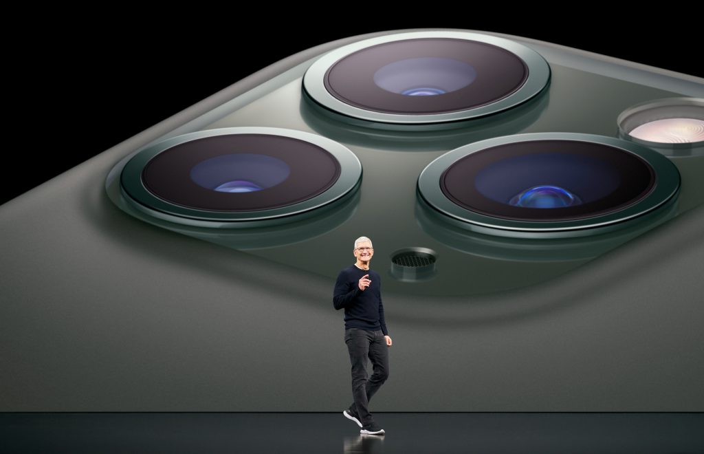 A person wearing glasses and a black shirt stands on a stage, gesturing with one hand. Behind them is an enormous image of Apple's green iPhone's camera array, highlighting the three camera lenses and flash, perhaps subtly referencing the Department of Justice's ongoing legal battle with tech giants.