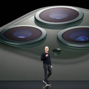 A person wearing glasses and a black shirt stands on a stage, gesturing with one hand. Behind them is an enormous image of Apple's green iPhone's camera array, highlighting the three camera lenses and flash, perhaps subtly referencing the Department of Justice's ongoing legal battle with tech giants.