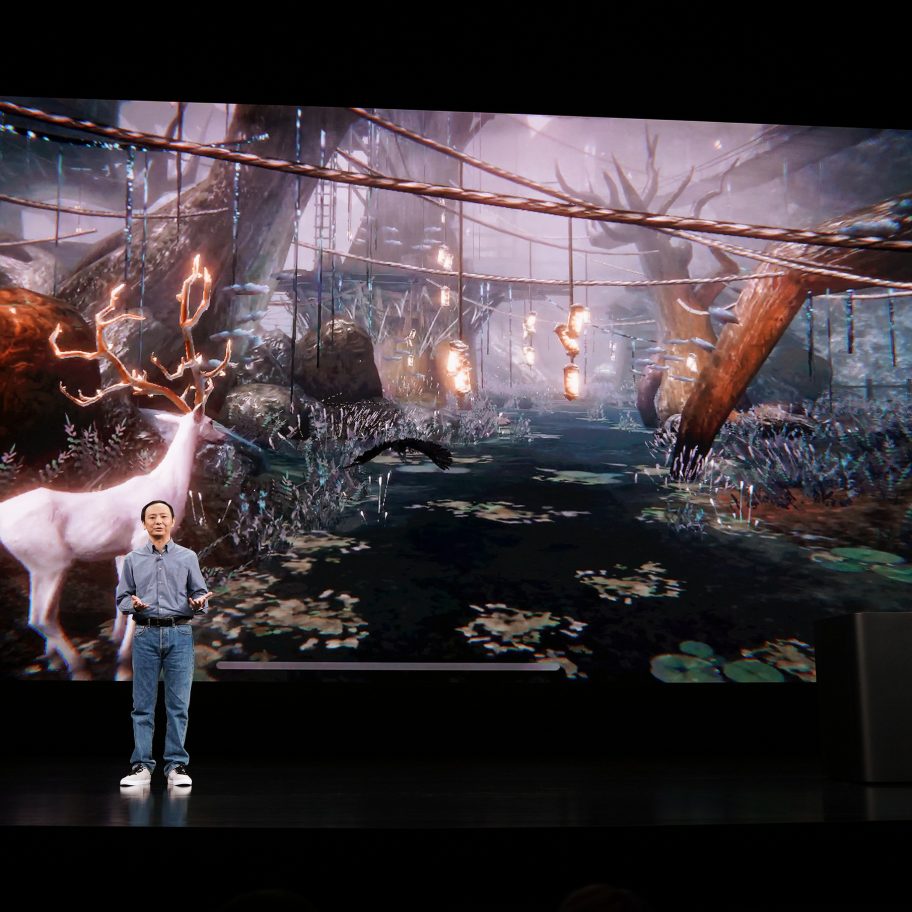 Two individuals present a video game on a large screen in a dark room. The screen displays a mystical forest scene with a glowing white deer and hanging lanterns. One person stands center stage speaking, while the other operates a device at a podium on the right.