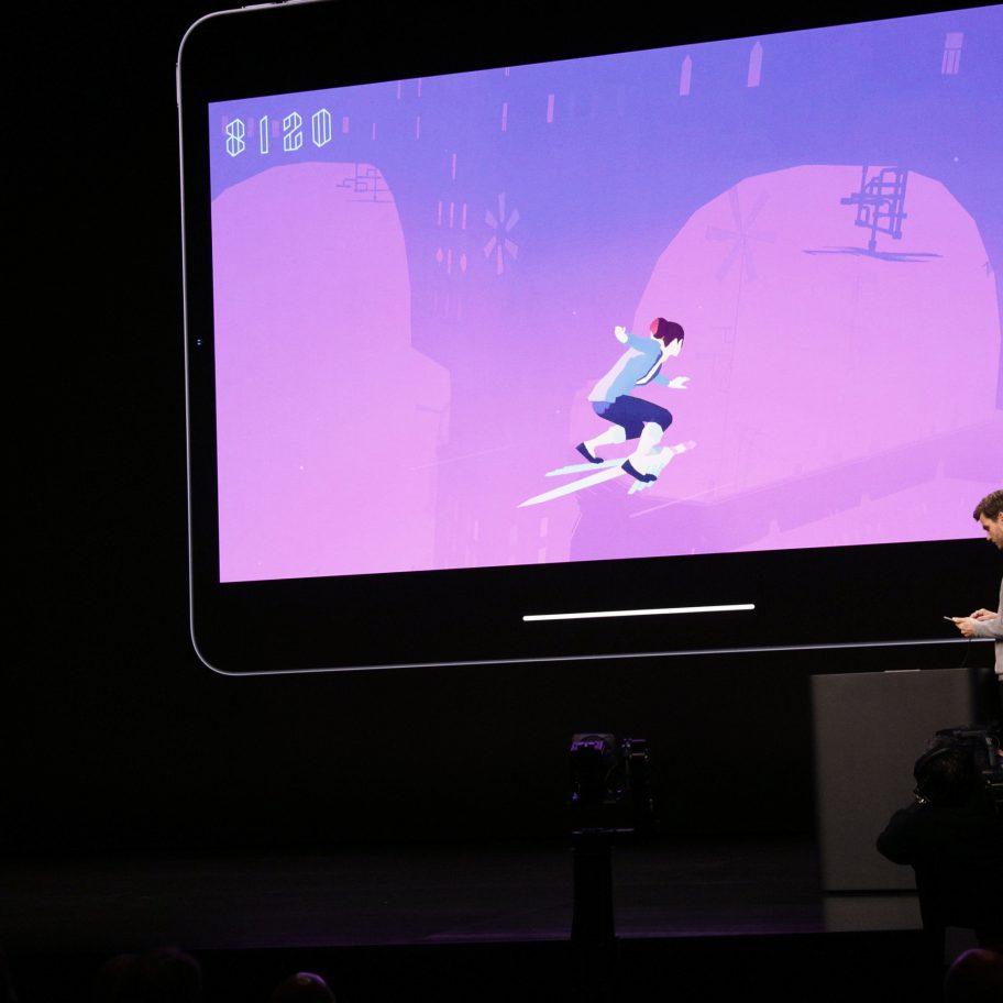 Two individuals stand on a stage presenting a large screen displaying a purple-toned animated game. The person on the left is speaking into a microphone, while the person on the right is using a device, possibly to control the game.