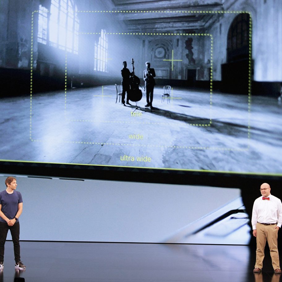 Two men stand on a stage beside a large screen displaying a photo-editing interface. The screen shows a black-and-white image of a spacious, sunlit room with three figures and a cello. The stage presentation appears to be about mobile photography features.