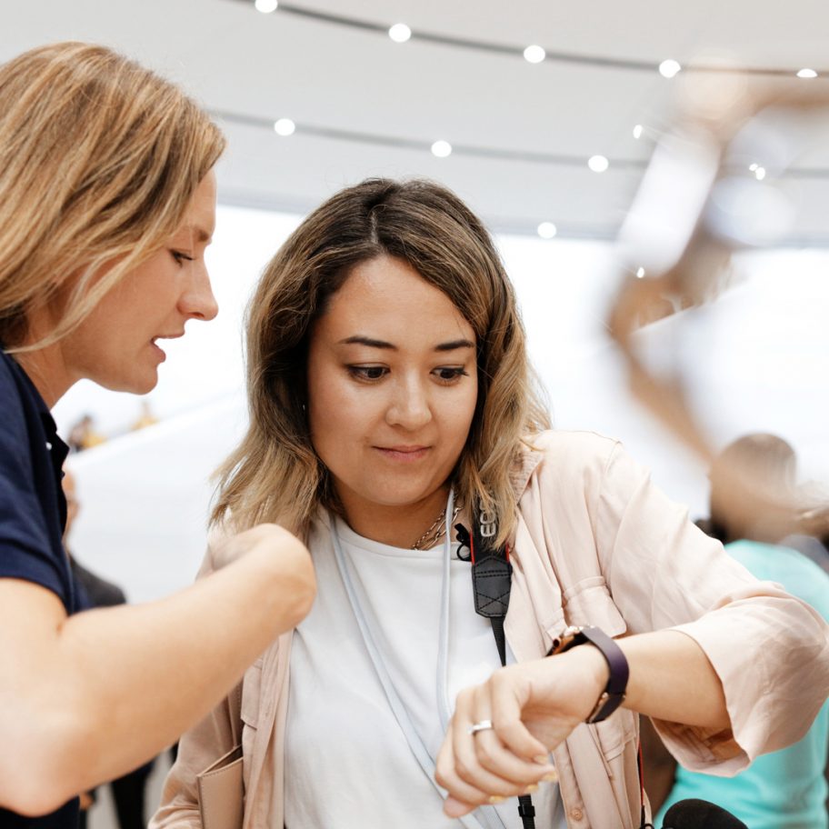 Two women are examining a smartwatch in a brightly lit indoor setting. Both have light brown hair and casual attire; one wears a navy shirt while the other has a white shirt with a beige jacket. Other people and a circular light fixture are visible in the blurred background.