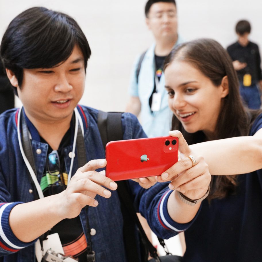 Two people are looking at a red smartphone together, with one person pointing at the screen. They appear to be engaged in a conversation. Several other individuals are in the background, out of focus. The scene appears to be indoors, possibly at an event or gathering.