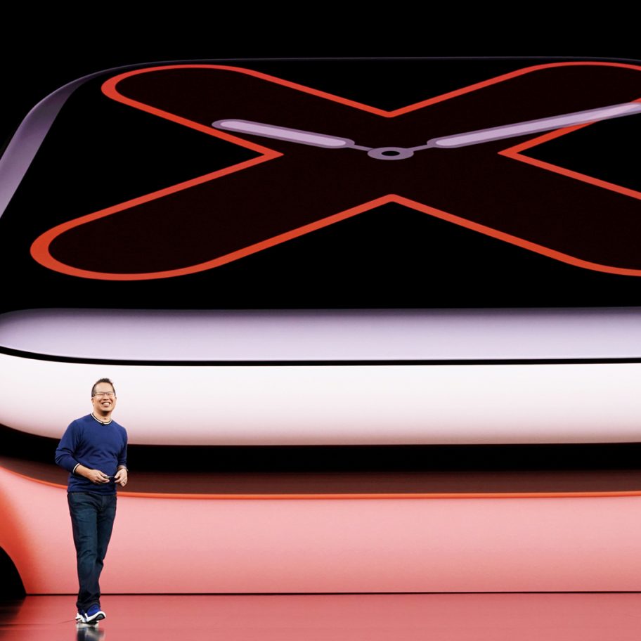 A person standing on a stage next to a large screen displaying a close-up of a smartwatch. The smartwatch has a minimalistic clock face with a large "X" indicating roman numeral ten. The person appears to be presenting or speaking about the product.