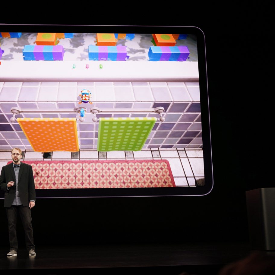 Two individuals stand on stage demonstrating a video game on a large screen behind them. One person speaks into a microphone while the other operates a device on a podium. The screen displays a colorful game with a character navigating a vibrant environment.