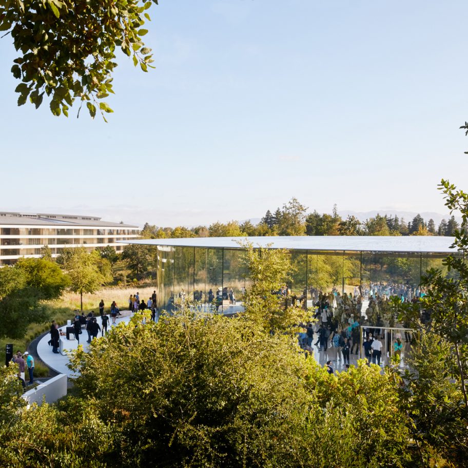 A large, modern glass building with a flat roof sits in the middle of a green, lush landscape. Many people can be seen inside the building, which is surrounded by trees and vegetation. Another building is visible in the background through the trees.