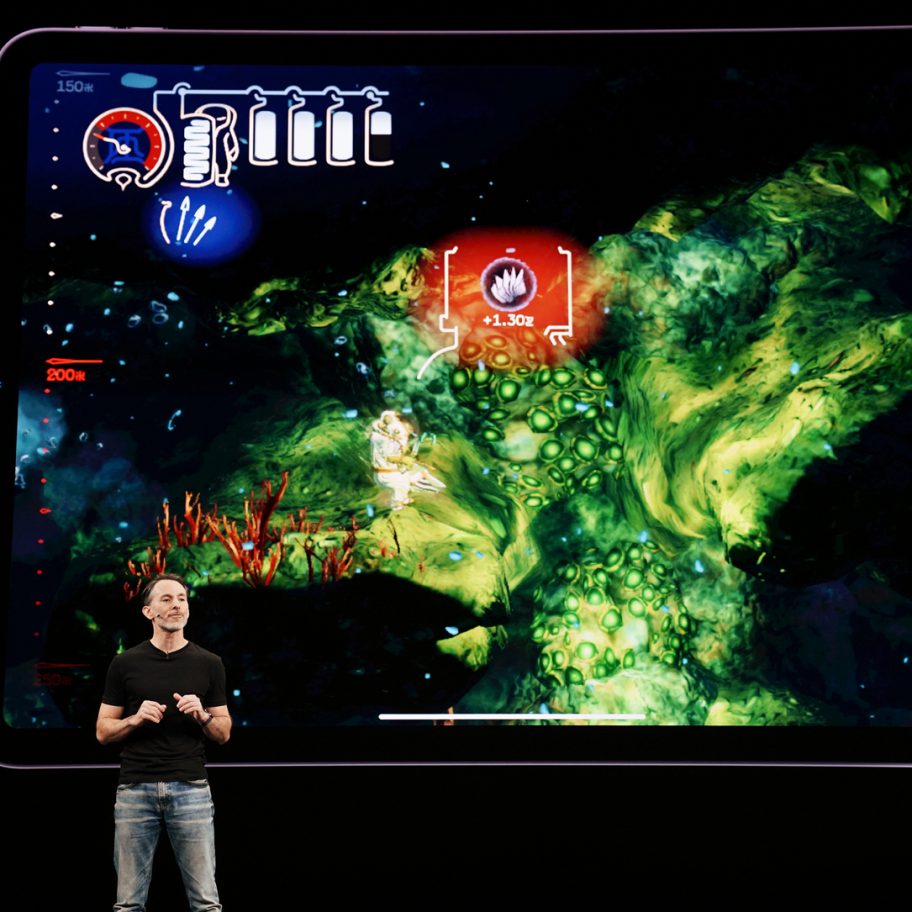 A presenter stands on stage holding a device in front of a large screen displaying a colorful underwater-themed video game interface. The background features intricate details of underwater flora and fauna along with various game icons.