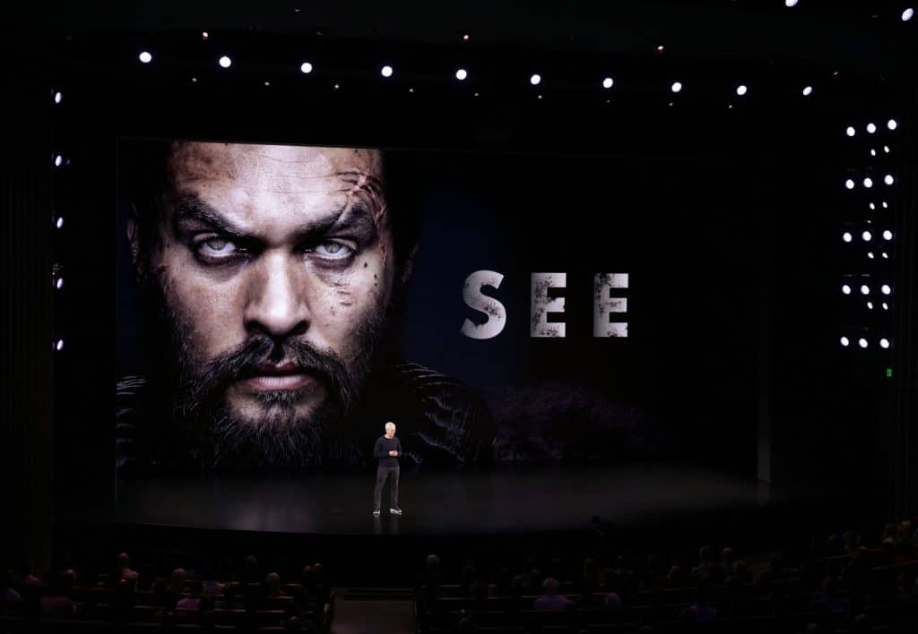 A person stands on a stage in a dark auditorium with a large screen behind them showing a close-up of a stern-faced individual and the word "SEE." The audience is seated in front, and stage lights illuminate the scene.