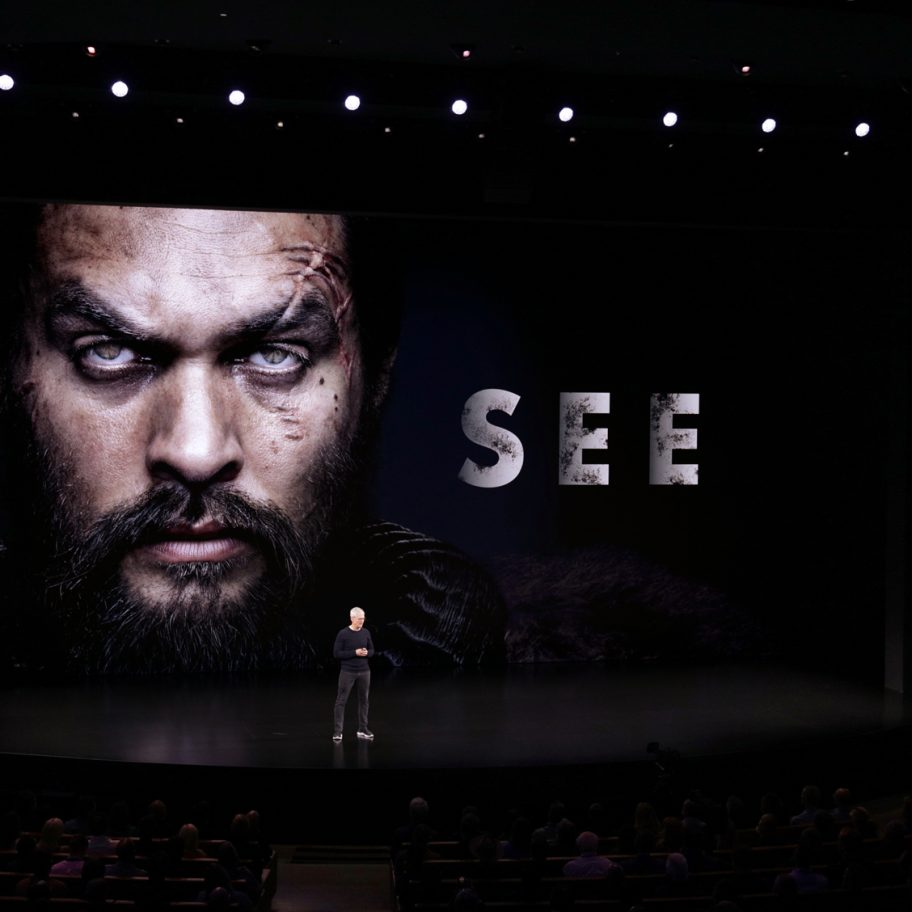 A person stands on a stage in a dark auditorium with a large screen behind them showing a close-up of a stern-faced individual and the word "SEE." The audience is seated in front, and stage lights illuminate the scene.