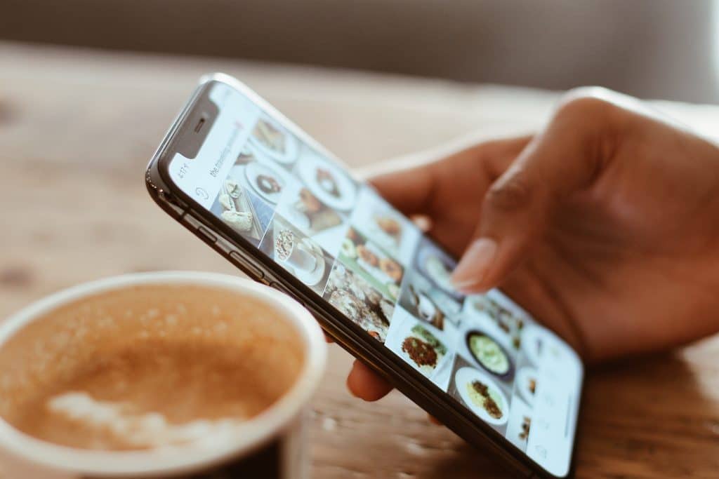 A hand holding an iPhone, swiping through a social media app displaying various food images. In the foreground, a cup of coffee with textured foam sits on a wooden table. The scene has a warm, cozy ambiance, with the iPhone's Touch ID subtly visible.