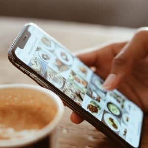 A hand holding an iPhone, swiping through a social media app displaying various food images. In the foreground, a cup of coffee with textured foam sits on a wooden table. The scene has a warm, cozy ambiance, with the iPhone's Touch ID subtly visible.