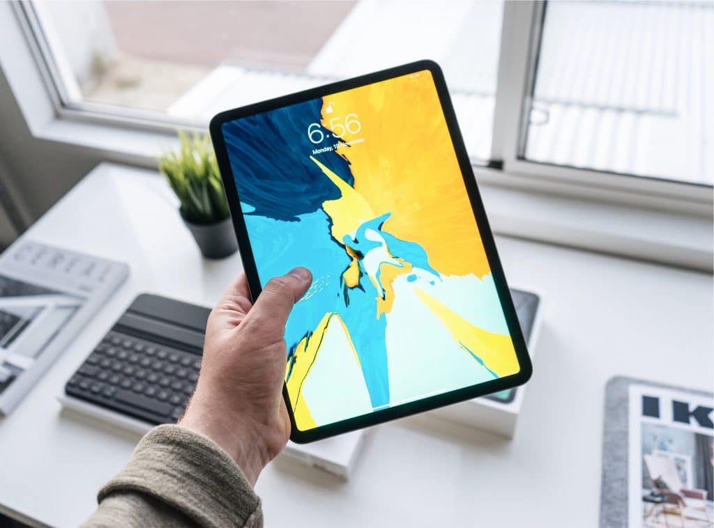 A hand holding an iPad with a colorful abstract display showing the time 6:56. The iPad is in an indoor setting near a window. On the table in the background, there is a MacBook, a small plant, and some papers.