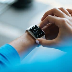 A close-up of a person's wrist wearing a smartwatch with a silver frame and black screen displaying the time, weather, and various app icons. The person uses their other hand to navigate the watch. The background is blurred, highlighting the focus on the watch that's subtly transforming industry with Apple healthcare features.