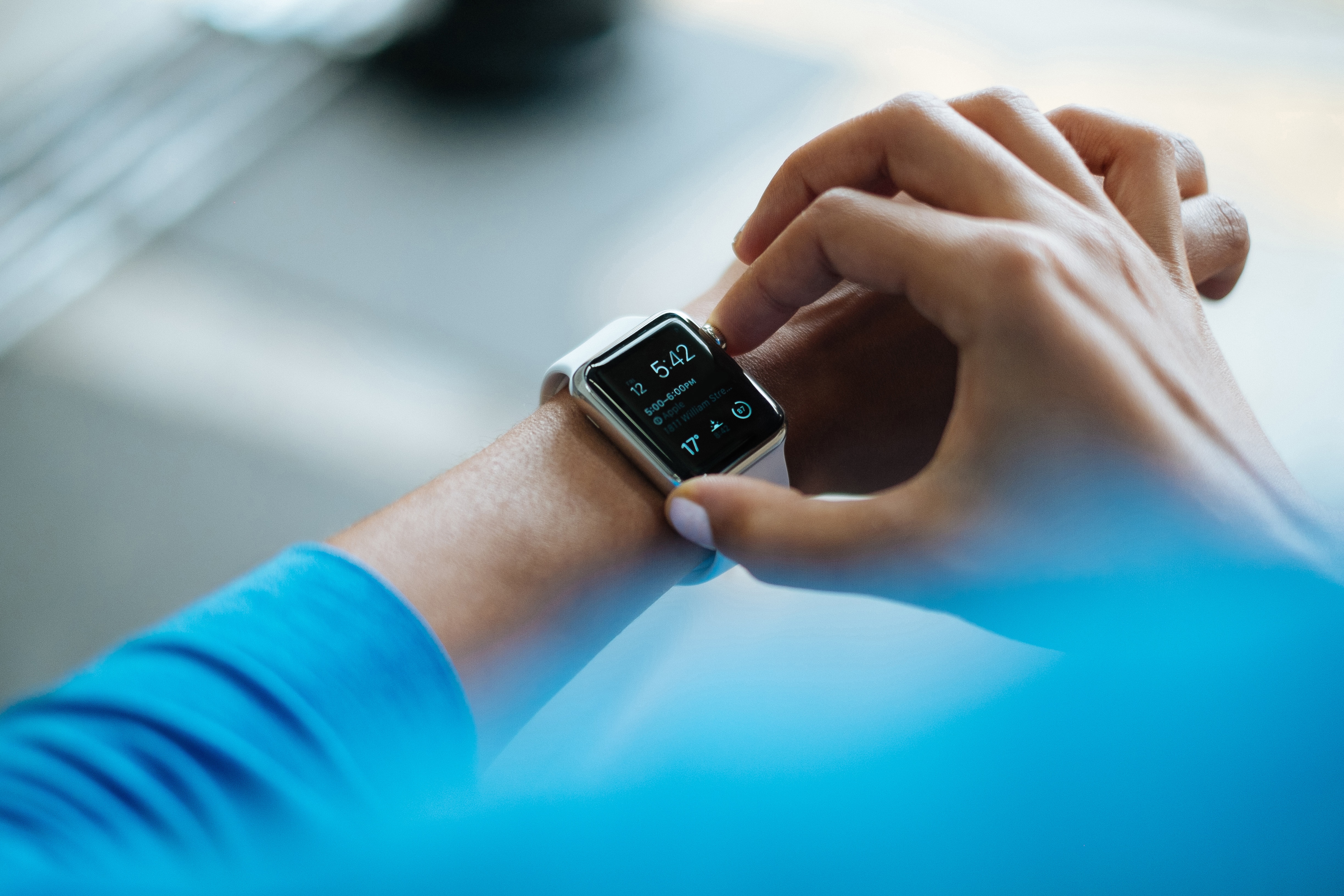 A close-up of a person's wrist wearing a smartwatch with a silver frame and black screen displaying the time, weather, and various app icons. The person uses their other hand to navigate the watch. The background is blurred, highlighting the focus on the watch that's subtly transforming industry with Apple healthcare features.