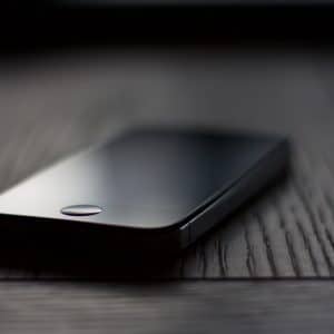 A black iPhone 4 with a reflective screen lies face up on a dark wooden surface. The image is taken with a shallow depth of field, causing the background to be blurred. The phone's power button and metal frame are clearly visible on its side.