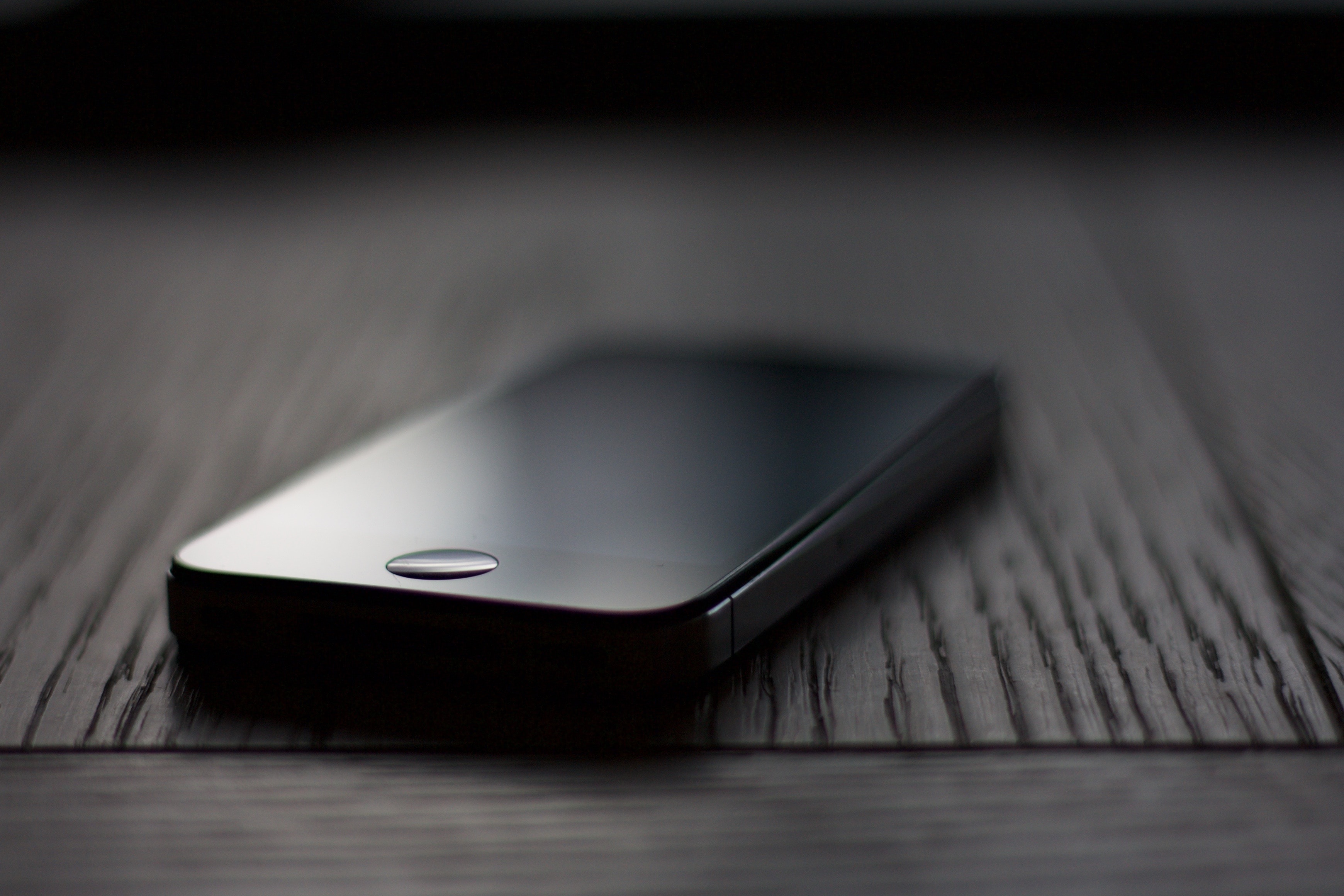 A black iPhone 4 with a reflective screen lies face up on a dark wooden surface. The image is taken with a shallow depth of field, causing the background to be blurred. The phone's power button and metal frame are clearly visible on its side.