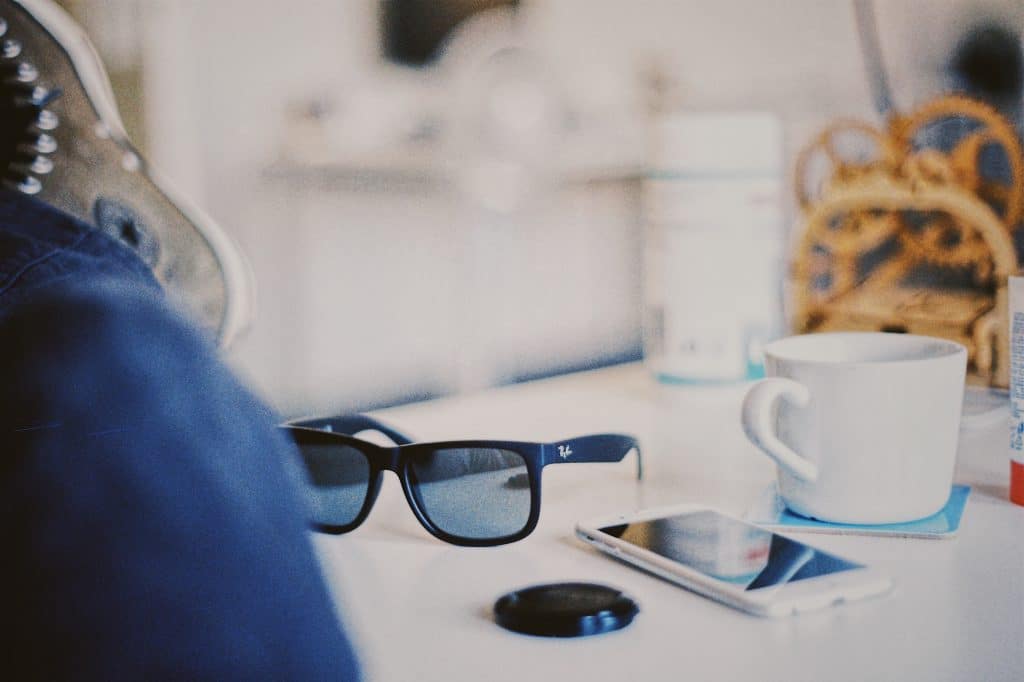 A close-up of a white table with a pair of smart glasses, a smartphone, a camera lens cap, and a white mug. In the background, there's a blurry set of pretzels and a cylindrical container. The overall scene appears casual and relaxed, reminiscent of everyday moments shared on Facebook in 2023.