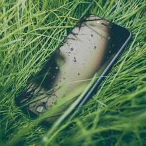 An Apple smartphone lies face-up in the middle of lush green grass. The screen, speckled with droplets of water, reflects the surrounding grass blades. The scene suggests it was recently raining or the phone was spilled on, embodying Apple's vision for greener products in harmony with nature.