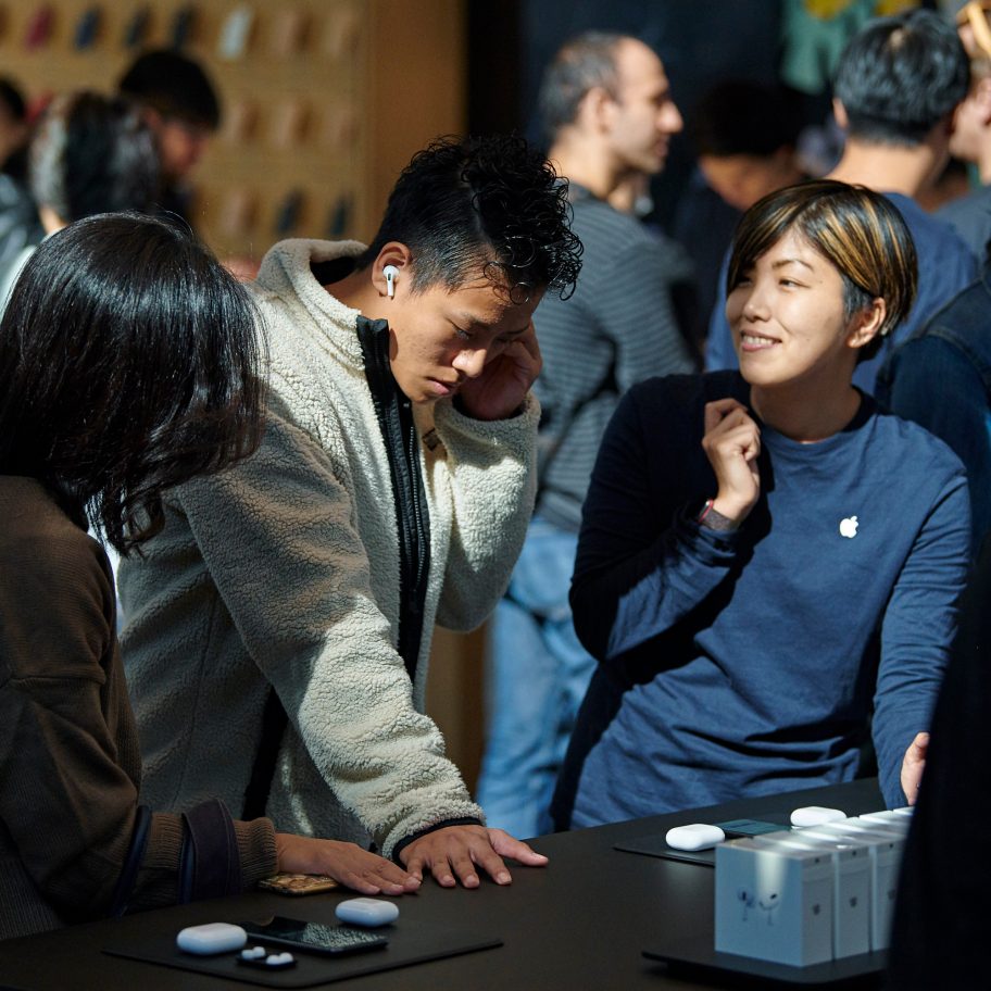 A group of people stands around a display table in a busy store. One person is wearing a beige jacket and trying on AirPods Pro, while another, in a navy shirt with an Apple logo, is smiling and looking towards the person trying the earbuds. The background is filled with more people and products on display.