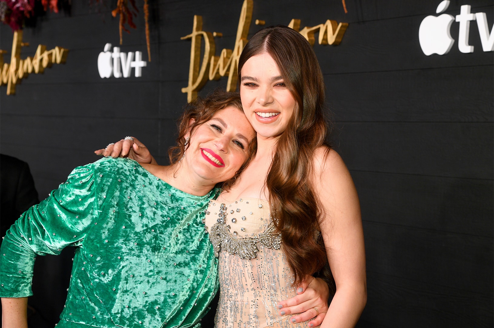 Two women pose together at an event at St. Ann’s Warehouse in Brooklyn, New York. The woman on the left wears a green velvet dress and has red lipstick, smiling with her arm around the shoulders of the woman on the right, who has long, wavy hair and is dressed in an elegant, embellished gown.