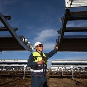 A person wearing a hard hat, safety vest, and sunglasses is inspecting solar panel installations at an outdoor solar farm under a clear sky. Holding documents or a tablet, they look up at the panels, ensuring everything aligns with the Green Supply Chain standards.
