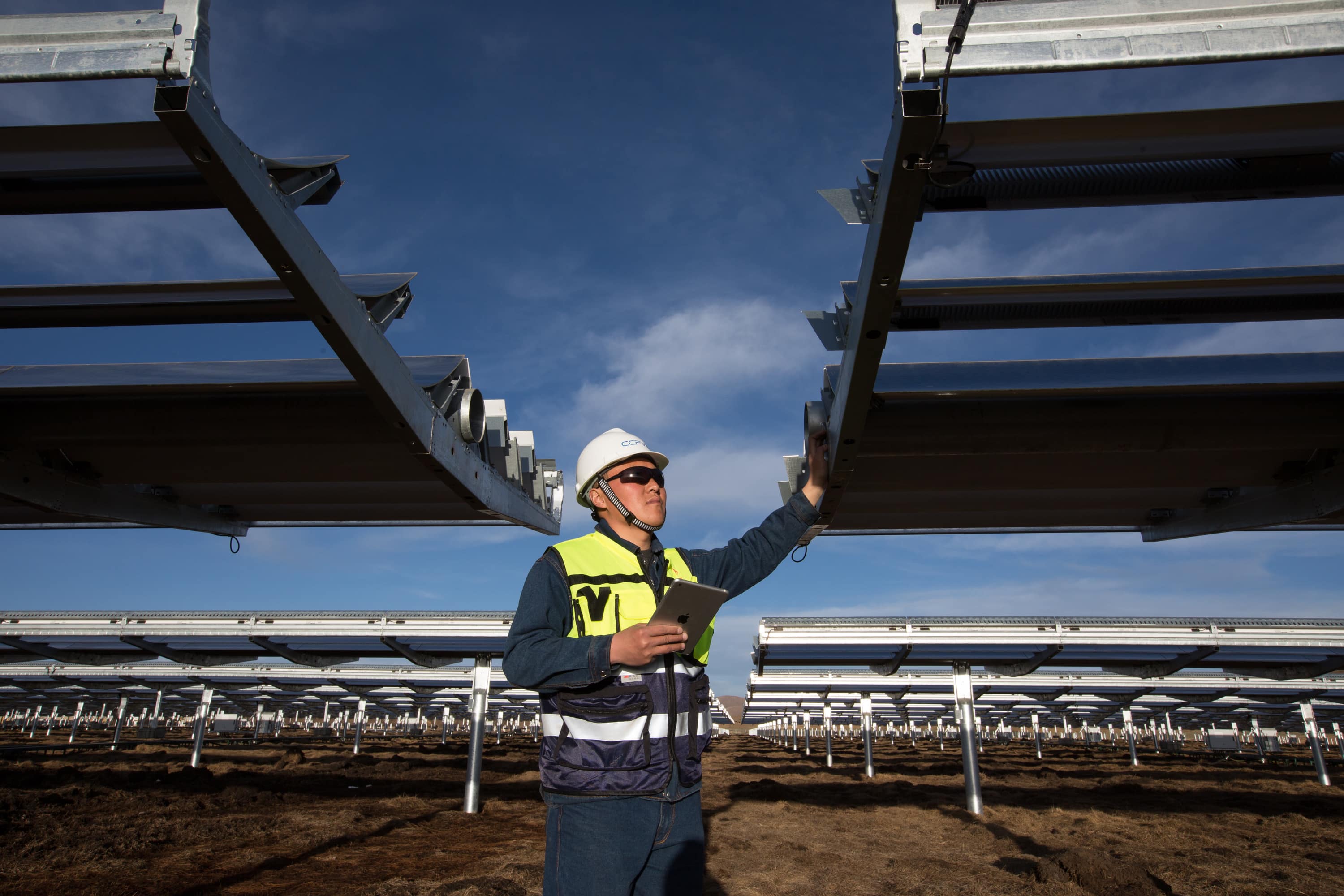 A person wearing a hard hat, safety vest, and sunglasses is inspecting solar panel installations at an outdoor solar farm under a clear sky. Holding documents or a tablet, they look up at the panels, ensuring everything aligns with the Green Supply Chain standards.