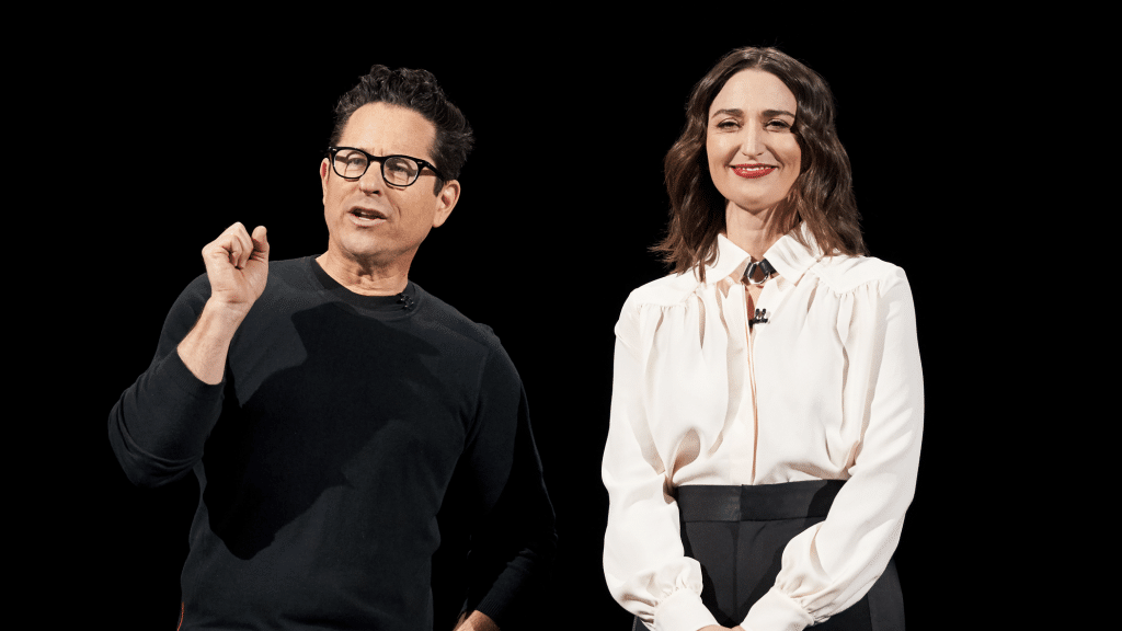 A man and a woman are standing side by side against a black background. The man, wearing glasses and a black shirt, is gesturing with one hand. The woman, in a white blouse and black pants, is smiling and looking ahead—just like in popular Apple TV+ shows.