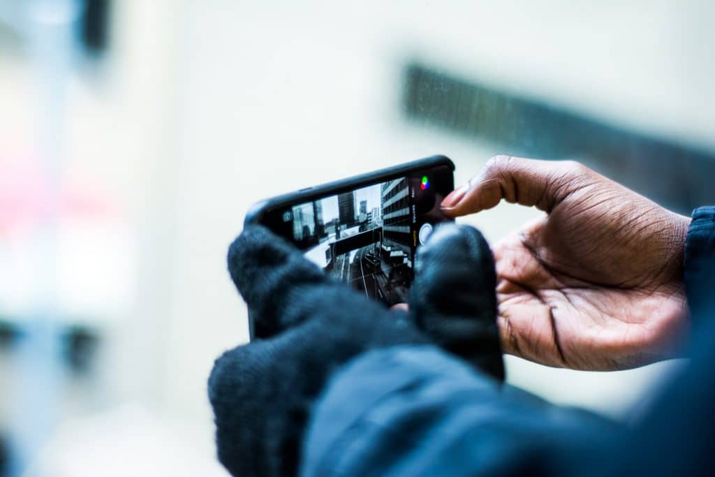 A person wearing black gloves is holding an iPhone SE 2 horizontally, capturing a photo of a street scene through a window. The screen of the smartphone displays a portion of the scene with buildings and light reflections, showcasing its vivid LCD display.