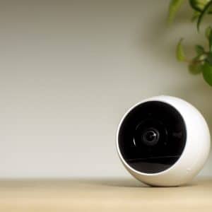 A white spherical security camera with a black lens sits on a light-colored wooden surface. A green leafy plant in a white pot with a wooden stand is visible in the background to the right of the camera. The background wall is plain and light-colored, evoking an aesthetic reminiscent of an Apple Store display for HomeKit Secure Video devices.