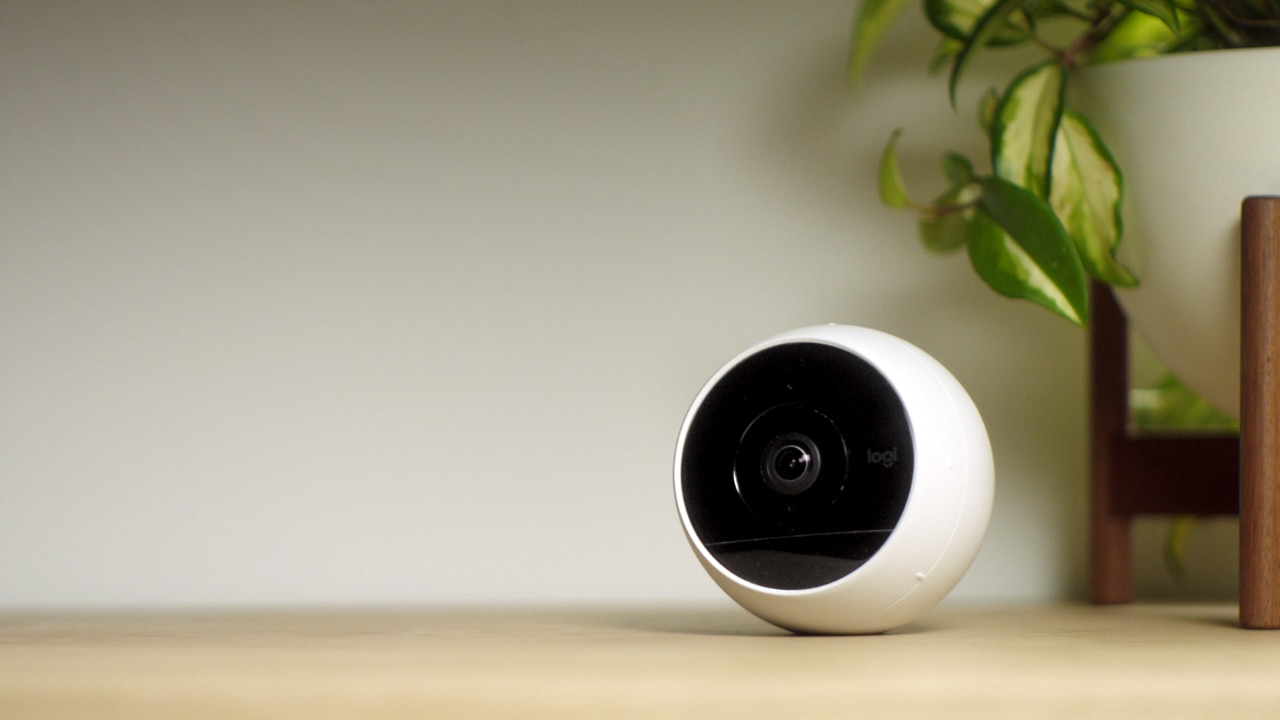 A white spherical security camera with a black lens sits on a light-colored wooden surface. A green leafy plant in a white pot with a wooden stand is visible in the background to the right of the camera. The background wall is plain and light-colored, evoking an aesthetic reminiscent of an Apple Store display for HomeKit Secure Video devices.