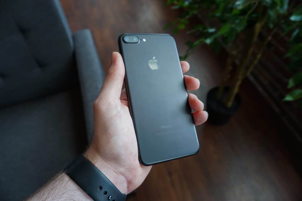 A hand holding a black iPhone SE 2, priced at $399, with the Apple logo on the rear. The background features a potted plant, a sofa, and wooden flooring, creating a cozy indoor setting. The person is wearing a black wristband on their left wrist.