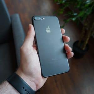 A hand holding a black iPhone SE 2, priced at $399, with the Apple logo on the rear. The background features a potted plant, a sofa, and wooden flooring, creating a cozy indoor setting. The person is wearing a black wristband on their left wrist.