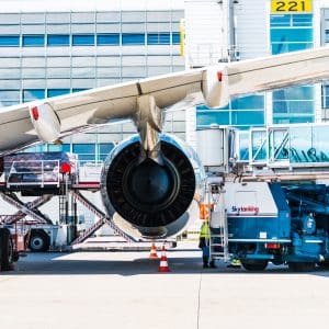 A close-up view of a jet engine of a parked airplane at San Francisco International Airport. Ground service vehicles are positioned around, with personnel likely assisting in maintenance or loading. The modern terminal design and boarding bridges are visible in the background.