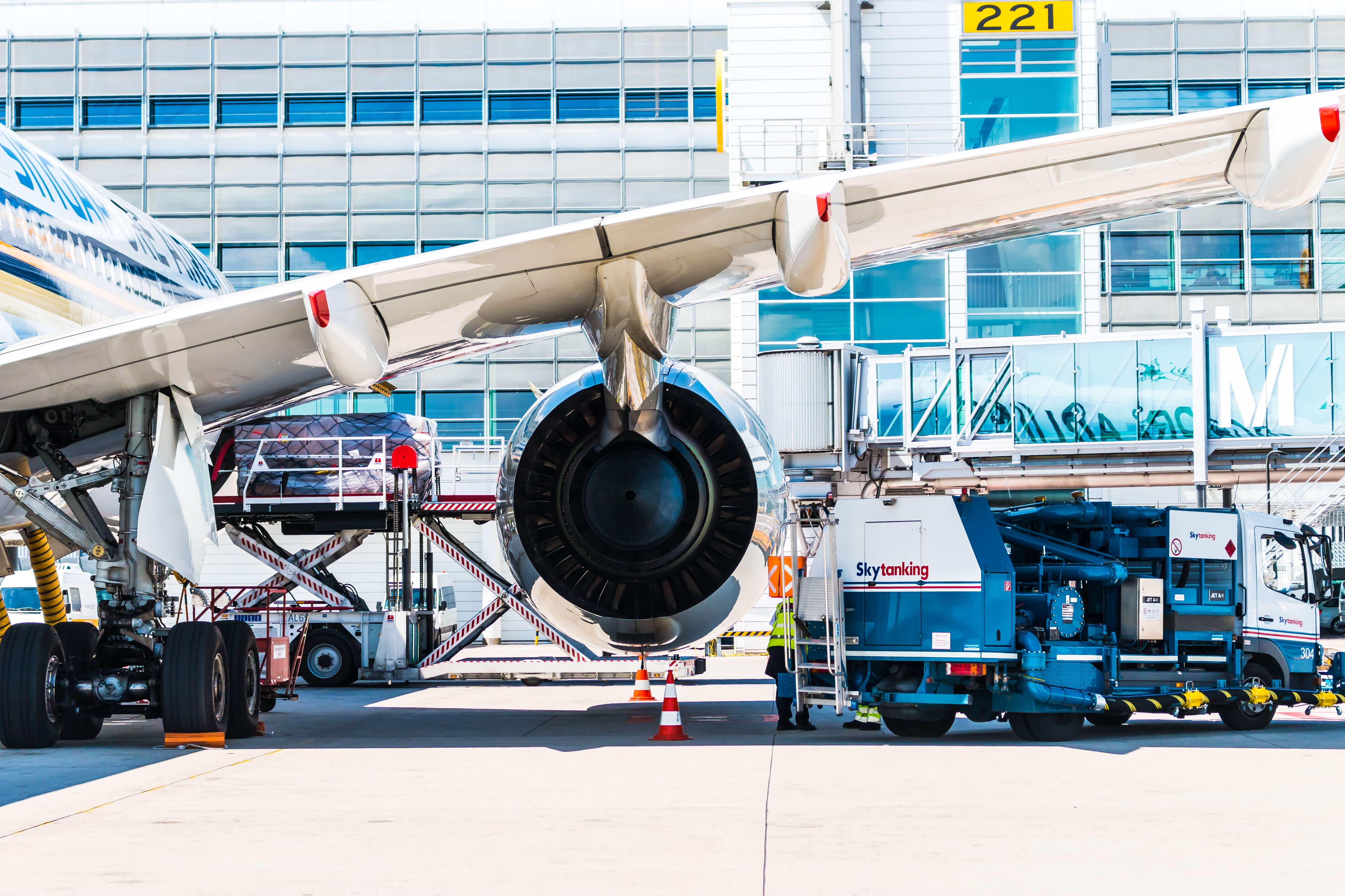 A close-up view of a jet engine of a parked airplane at San Francisco International Airport. Ground service vehicles are positioned around, with personnel likely assisting in maintenance or loading. The modern terminal design and boarding bridges are visible in the background.