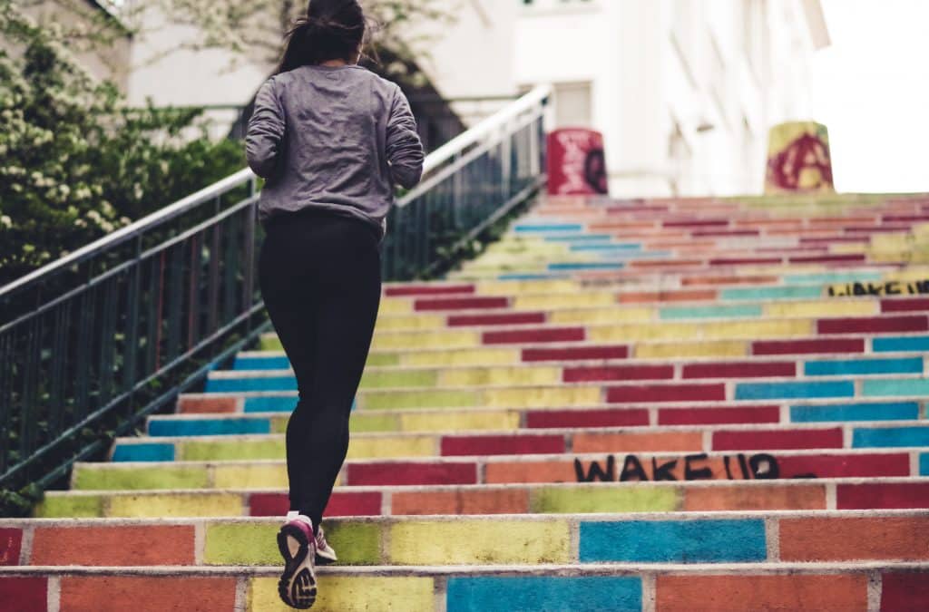 A person with a ponytail, wearing a gray jacket and black pants, is jogging up colorful, graffiti-covered stairs outside. The steps are painted in various bright colors with some words and designs visible. This vibrant scene highlights the joy of fitness and maintaining good health.