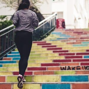 A person with a ponytail, wearing a gray jacket and black pants, is jogging up colorful, graffiti-covered stairs outside. The steps are painted in various bright colors with some words and designs visible. This vibrant scene highlights the joy of fitness and maintaining good health.