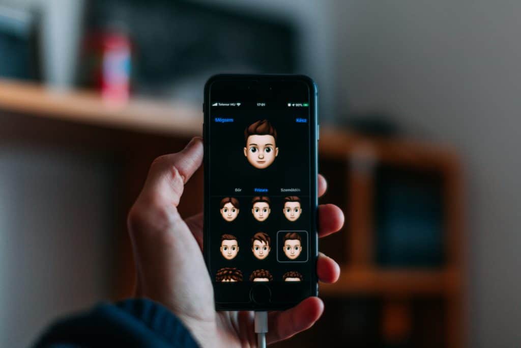 A hand holding an iPhone with a screen displaying an animated character creation app. The app, optimized for iOS 13, shows various customization options for hairstyles and facial features under the character’s head. The background is blurred, focusing on the phone's screen.