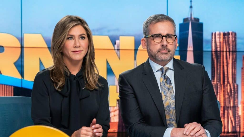 Two people sit in front of a colorful backdrop with large, partially visible letters. The person on the left has long, brown hair and is wearing a black outfit. The person on the right, possibly discussing recent negative reviews of "The Morning Show," has short, gray hair, glasses, and is wearing a suit with a patterned tie.