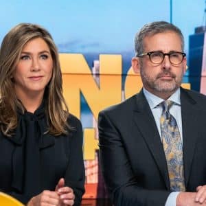 Two people sit in front of a colorful backdrop with large, partially visible letters. The person on the left has long, brown hair and is wearing a black outfit. The person on the right, possibly discussing recent negative reviews of "The Morning Show," has short, gray hair, glasses, and is wearing a suit with a patterned tie.