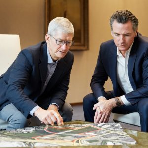 Two men in business attire are sitting and leaning over a large map on a table. One of the men is pointing at the map while the other attentively looks on, possibly discussing strategies for addressing the California housing crisis. The background features a plain wall and part of a U.S. flag is visible on the right side.