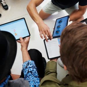 Four people sitting in a circle, each holding a tablet, presumably part of Apple's Everyone Can Code initiative. They appear to be collaborating or discussing something displayed on their screens. The image is taken from an overhead angle, showing the devices but not focusing on individual faces.