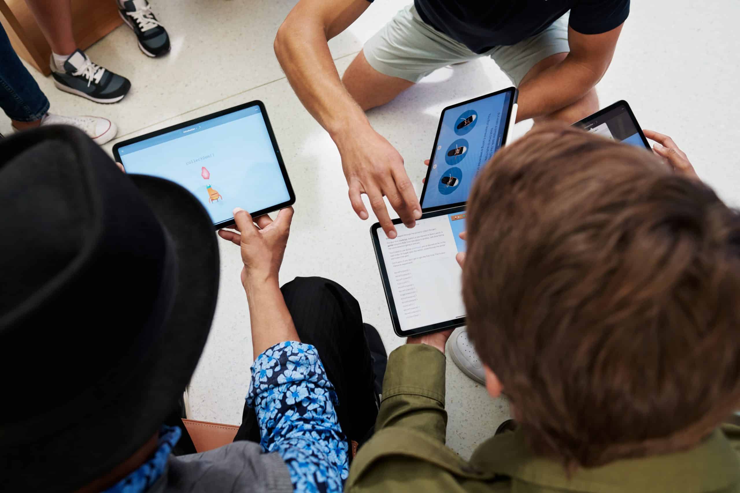 Four people sitting in a circle, each holding a tablet, presumably part of Apple's Everyone Can Code initiative. They appear to be collaborating or discussing something displayed on their screens. The image is taken from an overhead angle, showing the devices but not focusing on individual faces.