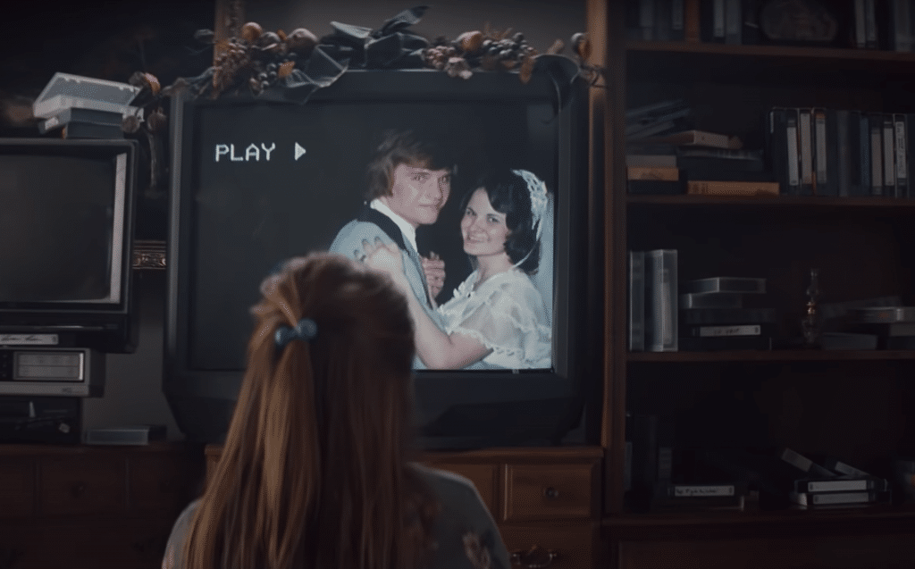 A person with a blue hair tie watches a video on an old television showing a bride and groom smiling and dancing. The TV, surrounded by books and VHS tapes, has a garland draped over the top of the screen. The word "PLAY" is displayed, reminiscent of "The Surprise," the heartwarming 2019 Holiday advert by Apple.