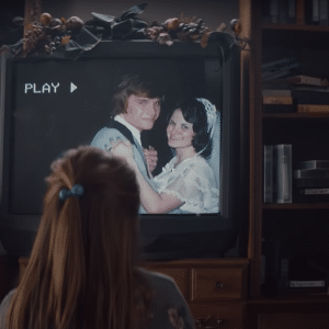 A person with a blue hair tie watches a video on an old television showing a bride and groom smiling and dancing. The TV, surrounded by books and VHS tapes, has a garland draped over the top of the screen. The word "PLAY" is displayed, reminiscent of "The Surprise," the heartwarming 2019 Holiday advert by Apple.