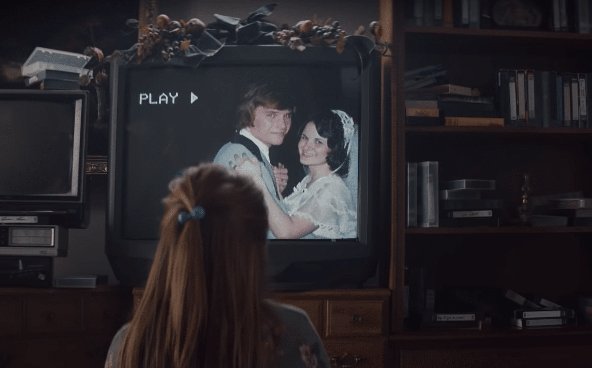 A person with a blue hair tie watches a video on an old television showing a bride and groom smiling and dancing. The TV, surrounded by books and VHS tapes, has a garland draped over the top of the screen. The word "PLAY" is displayed, reminiscent of "The Surprise," the heartwarming 2019 Holiday advert by Apple.