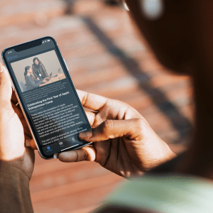 A person looks at a smartphone displaying an online news article. The article features an image of three people sitting on a couch. The person's finger hovers over the screen, perhaps about to scroll or tap through the Apple Developer App for updates from WWDC. The background is blurred.