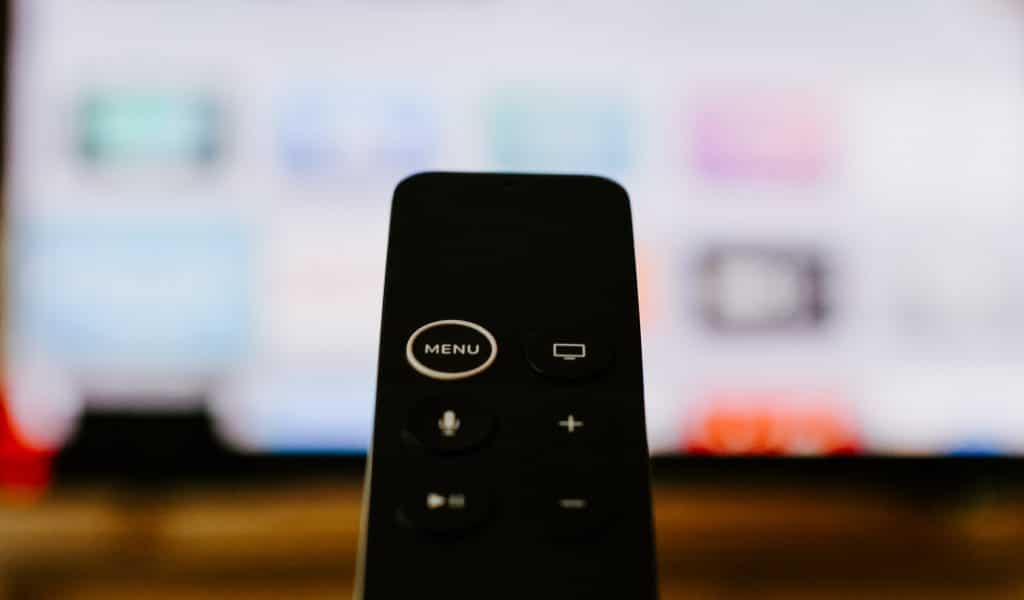 Close-up of a black remote control pointing towards a blurred TV screen in the background. The remote, designed for Apple TV, features buttons including "Menu," a screen icon, voice control, plus and minus symbols for volume, and play/pause. Perfect for Apple customers in the US.