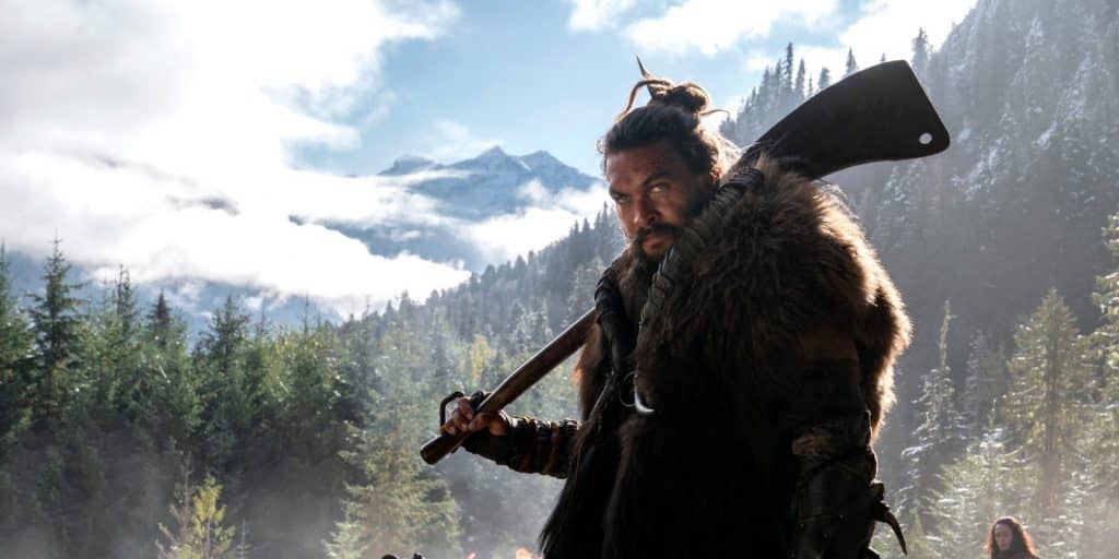 A man with long hair in a top knot, wearing fur clothing, stands in a misty forest with snow-covered mountains in the background. He holds a large axe over his shoulder, looking intently towards the camera, as if ready to step into a scene from Apple TV+'s epic series See.