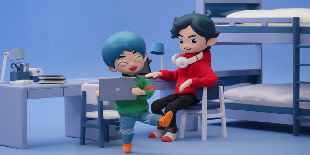 Two animated characters in a room with a bunk bed. One, with blue hair, wearing a green sweater, holds a tablet while sitting at a desk. The other, with black hair and a red hoodie, sits nearby with white headphones around their neck. They’re excitedly discussing the new additions to Apple Arcade's 100 games library.